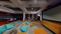 Located at 431 El Camino Real in Santa Clara, California, this single-floor amenity space is designed to serve as a dynamic hub for entertainment and social interaction. The centerpiece of the room is a large wall-mounted projection screen paired with a ceiling-mounted projector, creating an immersive viewing experience. Generous sectional sofas in gray, accented with teal and orange throw pillows, anchor the lounge area and provide ample comfortable seating for groups. A geometric patterned carpet in orange and gray adds a bold, modern aesthetic to the floor.

The space is thoughtfully zoned to accommodate multiple activities simultaneously. A raised built-in bench seating area with warm wood detailing offers an intimate nook, complemented by bistro-style tables and contemporary bar chairs positioned along the perimeter — ideal for casual dining or individual work. Recessed coffered ceilings with structured tray detailing add architectural depth, while strategic overhead and accent lighting sets a warm, inviting ambiance throughout.

An adjacent kitchenette area features light wood cabinetry, a gray countertop, and ample storage, providing convenient utility support for events and gatherings. The overall design blends contemporary finishes — rich wood tones, dark acoustic panels framed in walnut, and sleek modern furnishings — into a cohesive, energetic environment well-suited for screenings, social events, or collaborative use.