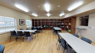 The Craft Room is a generously sized, single-floor space thoughtfully designed to support a wide range of creative and group activities. Light hardwood flooring runs throughout, lending the room a warm, clean aesthetic that complements the neutral beige walls. Overhead, a series of flush-mounted fluorescent light fixtures provide consistent, even illumination across the entire space.

One wall is lined floor-to-ceiling with built-in wooden shelving units in a warm walnut finish, offering ample storage for craft supplies, bins, and materials. Multiple large rectangular tables with black chairs are arranged throughout the open floor plan, providing generous workspace for individual or collaborative projects.

At one end of the room, a functional utility area includes warm-toned wooden upper and lower cabinetry along with a sink and continuous countertop — well-suited for cleanup and project preparation. A wall-mounted flat-screen television is also present in this area. Natural light filters in through windows along one wall, and an ironing board is available for textile-focused projects. The room's open layout and abundant storage make it a well-equipped, versatile space for a variety of hands-on creative pursuits.