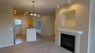 This single-floor space is designed with an open-concept layout that seamlessly connects the living and kitchen areas. The living room is anchored by a classic white-mantled fireplace with a black insert, complemented by a recessed niche above, ideal for displaying décor. Track lighting along the ceiling provides focused illumination throughout the space, while a decorative pendant chandelier serves as a focal point over the kitchen peninsula.

The kitchen features warm honey-toned cabinetry, a functional peninsula with an integrated sink, and countertops with a neutral finish. The peninsula offers casual seating potential and serves as a natural divider between the kitchen and living areas. Recessed lighting supplements the pendant fixture, ensuring the kitchen remains well-lit.

Soft wall-to-wall carpeting in the living area transitions to tile flooring in the kitchen, providing both comfort and practicality. The warm, cream-toned walls throughout create a cohesive and inviting ambiance. A hallway visible from the main living space suggests additional rooms beyond, contributing to the overall functional flow of this thoughtfully designed layout.