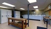 This single-floor laundromat offers a spacious, open layout designed to accommodate a high volume of customers efficiently. The facility is outfitted with a long row of commercial-grade top-loading washing machines and dryers lining the perimeter walls, providing ample capacity for simultaneous use. A bank of white storage lockers is positioned along one wall, offering customers a secure place to store personal belongings while doing laundry. At the center of the room stands a large, sturdy wooden folding table with bench seating, giving patrons a practical surface for sorting and folding laundry. The floor is finished in light-colored tile, contributing to a clean and easy-to-maintain environment. Overhead, a drop ceiling with recessed fluorescent panel lighting ensures the space is evenly and brightly lit throughout. Dark structural support columns are positioned throughout the floor, adding an industrial character to the overall aesthetic. The walls are painted in a muted olive green tone, providing a neutral backdrop that complements the functional design. Overall, this laundromat is thoughtfully arranged to maximize usability, comfort, and efficiency for its patrons.