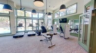 This fitness center is designed to offer a comprehensive workout experience within a spacious, single-level layout. The room's most striking architectural feature is its soaring ceiling height, which lends an open, airy quality reinforced by two stacked rows of large, green-trimmed windows along multiple walls. These expansive windows allow generous natural light to fill the space while providing views of the surrounding outdoor environment, creating an inviting atmosphere.

The interior design incorporates sage green wainscoting and window trim that gives the space a cohesive, polished aesthetic. Large pendant dome lights hang from the ceiling alongside a ceiling fan, ensuring both effective illumination and air circulation throughout the room. Full-length mirrors are mounted along one wall, visually expanding the space and providing functional feedback during workouts.

The fitness center is equipped with a well-rounded selection of exercise machines, including treadmills, a stair climber, and multiple strength-training machines. A weight rack with dumbbells is also available for free-weight exercises. A wall-mounted flat-screen television adds an entertainment option for users. The flooring consists of light-toned carpet, softening the space both visually and acoustically. Green-framed glass doors provide access to adjoining spaces, maintaining visual connectivity throughout.