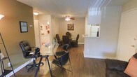 This single-floor space offers a practical and welcoming layout designed to accommodate both work and everyday living. The main area seamlessly blends a workspace and dining zone, unified by rich, dark hardwood-style flooring that runs throughout. A glass-top desk with rolling office chairs anchors the workspace portion of the room, while a round dining table surrounded by dark upholstered chairs serves as the dining area. A ceiling fan with an integrated light fixture provides both comfort and illumination overhead. A half-wall partition with a built-in shelf adds subtle architectural definition to the space without sacrificing openness, and a window fitted with a roller blind allows for natural light control. Decorative touches, including wall art, add a personalized warmth to the neutral, beige-toned walls. An open doorway leads into a bright, white kitchen visible in the background, enhancing the sense of flow and connectivity between areas. The overall ambiance is functional yet comfortable, making efficient use of the available space while maintaining a clean, organized aesthetic suitable for both professional and residential use.
