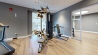 Located at 195 Dundas St in London, Ontario, this fitness facility offers a thoughtfully designed workout environment spread across two interconnected areas. The gym features light hardwood flooring throughout, neutral grey and blue-toned walls, and recessed lighting that creates a clean, modern aesthetic.

The main workout area is generously proportioned, accommodating a variety of cardio equipment including treadmills, a stationary bike, and an elliptical trainer. Large windows along the perimeter flood the space with natural light while offering views of the surrounding urban streetscape. A water fountain station is conveniently positioned near the entrance, alongside a frosted glass door that provides easy access.

A dedicated strength training zone houses a multi-function cable machine and weight bench, positioned adjacent to a floor-to-ceiling window that further enhances the sense of openness. A separate alcove provides space for a full dumbbell rack with a range of free weights and kettlebells, neatly organized for easy access.

Additional features include exercise mats for stretching and floor-based workouts, recessed spotlights, and white trim accents that complement the contemporary finish throughout. The layout is practical and efficient, offering a complete fitness experience within a compact yet functional footprint.
