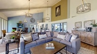 Located at 3516 Coronado Dr in Sarasota, Florida, this single-story community facility offers an impressive blend of casual elegance and practical amenities. The centerpiece of the space is a sweeping great room defined by soaring vaulted ceilings with exposed beams, warm hardwood-style flooring, and a soft coastal color palette of blues and golds. Three statement cage-style chandeliers hang dramatically from the ceiling, casting warm light over a generously sized sectional seating area anchored by a rustic barn-door media console. A floor-to-ceiling wall of windows along one side floods the interior with natural light and frames lush, mature greenery outside. A dedicated dining area with wicker seating complements the lounge space, while a workstation and intimate seating nook offer functional areas for residents. A large decorative clock and curated artwork add character throughout. Adjacent to the main lounge, a well-appointed fitness center is outfitted with cardio machines including treadmills and an elliptical, a full rack of free weights, a power rack, and cable machines, all set on durable tile carpet flooring with blue accent columns. Windows and glass doors in the gym provide views of the outdoor pool area, keeping the space bright and connected to the outdoors.