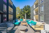 Located at 2520 College Ave in the heart of Berkeley, CA, this outdoor amenity space is a thoughtfully designed courtyard nestled within a contemporary multi-story residential building. The space is defined by its modern aesthetic, with a smooth concrete surface that extends throughout the open-air layout, providing a clean and functional foundation for communal living.

At the center of the courtyard, a generous lounge area features an arrangement of vibrant teal and blue outdoor sofas, chairs, and chaise lounges accented by warm wood coffee tables, creating an inviting setting for relaxation and social gatherings. The space receives abundant natural sunlight, enhancing its open and airy character.

A striking large-scale, colorful geometric mural adorns one of the building's exterior walls, serving as a bold artistic focal point that energizes the space with dynamic shapes and vivid hues. Adjacent to the mural, a stainless steel BBQ grill station offers a dedicated area for outdoor cooking and entertaining.

The courtyard is flanked on multiple sides by the building's gray stucco exterior with white-trimmed windows and private balconies overhead, creating an intimate yet expansive feel. A secondary open terrace area extends to one side, framed by mature trees and white planter boxes, offering a quieter retreat. The overall ambiance is lively, modern, and community-focused, ideal for residents seeking vibrant outdoor living in an urban setting.