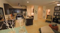This single-floor residence offers a thoughtfully designed open-concept layout that seamlessly connects the kitchen, dining, and living areas, creating a spacious and inviting atmosphere. The modern kitchen is a standout feature, showcasing dark espresso cabinetry that contrasts beautifully against warm beige walls and light flooring. Stainless steel appliances, including a gas range, built-in microwave, and a full-size refrigerator, complement the granite countertops and a central island that serves as both a prep space and a casual gathering point. Track lighting overhead illuminates the kitchen efficiently, while recessed lighting throughout the open area enhances the bright, airy ambiance. The light-toned flooring flows continuously across the space, reinforcing the sense of openness and cohesion. Decorative accents, including sculptural white branch arrangements and stone-filled display pieces, add an artistic touch to the interior. A hallway visible from the main living area leads to additional rooms, each accessed through white panel doors, providing a clean, classic finish. The overall design balances contemporary style with functional living, making this a well-appointed and visually cohesive space.