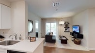 This single-floor residence offers a thoughtfully designed open-concept layout that seamlessly connects the kitchen and living areas, creating a spacious and cohesive living environment. The living room is anchored by a sleek dark sofa and a low-profile coffee table, both set atop a light-toned area rug that contrasts beautifully with the warm hardwood flooring running throughout the space. Natural light pours in through floor-to-ceiling sliding glass doors dressed with sheer white curtains, leading directly to an outdoor patio area with views of lush greenery beyond. The clean, light-toned walls and smooth white ceilings enhance the sense of openness and airiness throughout. A contemporary entertainment wall features floating dark shelves displaying curated décor alongside a flat-screen television. The adjacent kitchen area showcases a bright white countertop with a breakfast bar, offering casual seating and a natural transition between the cooking and living zones. A decorative round mirror adds a stylish focal point near the entryway, while carefully chosen accent lighting and wall-mounted fixtures contribute to the modern, refined ambiance of this well-appointed residence.