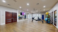 This cardio room in Annapolis, Maryland, offers a clean, well-appointed exercise space designed to support a variety of fitness activities. The room features warm hardwood flooring throughout, lending an inviting yet functional aesthetic to the space. Recessed lighting and dual ceiling fans ensure the environment remains bright and comfortable during workouts.

A standout feature of the room is the large mirrored wall panel, which visually expands the space and allows users to monitor their form during exercise. The mirrors are framed in crisp white trim, complementing the neutral, light-toned walls. A wall-mounted flat-screen TV is positioned for easy viewing during cardio sessions.

The equipment lineup includes a Concept2 rowing machine, stationary spin bikes, stability balls in multiple sizes, resistance bands, and a set of hand weights neatly organized on a low cubby-style storage shelf. A cork bulletin board with informational postings and a digital clock are mounted near the entry area, along with a hand sanitizer dispenser and a small waste bin.

Natural light flows in through a row of white-framed French doors along one wall, offering views of the surrounding outdoor greenery. Additional access is provided through rich, dark wood panel doors that add a touch of classic elegance. The overall ambiance is polished, practical, and welcoming for daily fitness use.