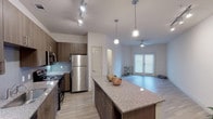 The Coral is a thoughtfully designed one-bedroom, one-bathroom residence that makes excellent use of its single-floor layout. The open-concept kitchen and living area serves as the heart of the unit, featuring rich dark-wood cabinetry, a subway tile backsplash, stainless steel appliances including a refrigerator, range, microwave, and dishwasher, and a large granite-topped island with pendant lighting overhead. Track lighting illuminates the kitchen, while the adjoining living area benefits from natural light through a set of windowed double doors. Light wood-style flooring flows throughout the main living spaces, lending a warm, cohesive feel. The bedroom is a comfortable, carpeted retreat with a ceiling fan, a window with blinds for natural light and privacy, and direct access to a walk-in closet. The bathroom is finished with classic white subway tile surrounding a full soaking tub and shower combination, complemented by tile flooring for a clean, bright aesthetic. Throughout the unit, neutral tones and modern fixtures create a contemporary yet welcoming atmosphere well-suited for everyday living.