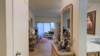This single-floor residence offers a richly appointed interior defined by classic elegance and carefully curated décor. The living area serves as the heart of the space, featuring plush carpeted flooring and an abundance of natural light streaming in through a large sliding glass door dressed with vertical blinds. The room is furnished with a graceful chaise lounge and accompanying seating, arranged to create an intimate and welcoming atmosphere. Ornate details are evident throughout, from the elaborately framed floor mirror near the entryway to the decorative glass-front display cabinet that showcases collectibles and keepsakes. Bronze sculptural figurines and artful accessories are thoughtfully placed on wicker and carved wood side tables, lending a gallery-like quality to the space. Framed artwork adorns the walls, adding depth and character to the interior. The entryway provides a graceful transition into the main living area, hinting at the layered and personal aesthetic carried throughout the home. Overall, the space presents a warm, collector's sensibility with a timeless, traditional charm that makes it feel both lived-in and distinctly refined.