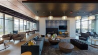 Located at 2109 S Wabash Ave in Chicago's South Loop neighborhood, this single-floor amenity space offers a thoughtfully designed environment that seamlessly blends comfort, functionality, and contemporary style. The interior is defined by a dynamic mix of ceiling treatments, including warm wood slat panels, dark painted sections, and exposed industrial elements, all contributing to a layered, modern aesthetic.

The main lounge area is anchored by a built-in gas fireplace set against a textured blue accent wall, flanked by a wall-mounted television and accented by recessed lighting above. Comfortable seating arrangements featuring tufted sofas, bold throw pillows, and sculptural rounded coffee tables create an inviting gathering space. A wet bar with wood cabinetry, a dark stone countertop, and subway tile backsplash provides convenient service access nearby.

A long communal work table with bar-height seating extends along the perimeter, bathed in abundant natural light streaming through floor-to-ceiling curtain wall glazing. These expansive windows frame views of the surrounding Chicago skyline and open directly to a landscaped outdoor terrace. The terrace, strung with Edison lights and furnished with seating areas, extends the usable space outdoors with open city views. A separate seating alcove with black leather chairs and brass-accented tables completes the interior, offering a more intimate setting within the larger open-plan layout.