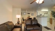 This single-floor residence offers a thoughtfully designed open-concept layout that seamlessly connects the living, dining, and kitchen areas. The living room is a comfortable and inviting space, furnished with rich dark leather sofas and accented by a decorative table lamp that casts a warm glow. A ceiling fan with an integrated light fixture overhead adds both functionality and a casual charm to the room.

The dining area sits just beyond the living room, featuring a round wooden table surrounded by dark chairs, creating an intimate setting for meals. Track lighting installed along the ceiling provides focused illumination throughout this shared space. Framed wall art and a small potted plant add tasteful decorative touches that enhance the overall ambiance.

The kitchen is accessible via a raised breakfast bar counter, which serves as a natural divider between the cooking and living spaces. White cabinetry lines the kitchen walls, and a microwave is visible along the upper shelving, indicating a well-equipped cooking area. The light-toned walls and neutral flooring throughout the home contribute to a bright and airy feel, making the space feel open and welcoming. Overall, the layout is practical and efficiently organized, suited for comfortable single-floor living.
