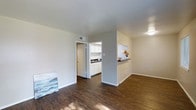 This single-floor space is characterized by its open, functional layout and consistent use of rich, dark hardwood flooring that runs throughout the main living area. The living space is bright and uncluttered, with white walls and a smooth white ceiling that together create a clean, neutral backdrop well-suited to a variety of furnishing styles. A flush-mounted ceiling light provides overhead illumination, complementing the natural light that filters into the space.

A partial wall with a pass-through counter separates the living area from the kitchen, maintaining an open feel while defining each zone. The kitchen features white cabinetry that extends to the upper walls, offering ample storage, along with a dark countertop that provides a subtle contrast. A sink is visible along the far wall, and the space is finished with the same consistent flooring. A doorway off the kitchen leads to an adjoining room, adding to the overall connectivity of the layout.

The overall ambiance is minimal and practical, with a neutral palette that keeps the space feeling open and easy to personalize.