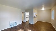 This single-floor space is characterized by its open, functional layout and consistent use of rich, dark hardwood flooring that runs throughout the main living area. The living space is bright and uncluttered, with white walls and a smooth white ceiling that together create a clean, neutral backdrop well-suited to a variety of furnishing styles. A flush-mounted ceiling light provides overhead illumination, complementing the natural light that filters into the space.

A partial wall with a pass-through counter separates the living area from the kitchen, maintaining an open feel while defining each zone. The kitchen features white cabinetry that extends to the upper walls, offering ample storage, along with a dark countertop that provides a subtle contrast. A sink is visible along the far wall, and the space is finished with the same consistent flooring. A doorway off the kitchen leads to an adjoining room, adding to the overall connectivity of the layout.

The overall ambiance is minimal and practical, with a neutral palette that keeps the space feeling open and easy to personalize.