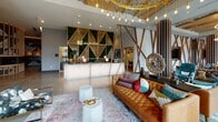 Located at 1500 Sherman Ave in Evanston, IL, this single-level lobby space at Albion Evanston makes an immediate impression with its bold, eclectic interior design. The expansive layout features polished concrete floors that transition into wide-plank reclaimed hardwood, creating a dynamic contrast underfoot. Soaring ceilings are adorned with exposed wood beam detailing and dramatic tiered gold ring chandeliers with Edison-style bulbs, lending the space a warm, upscale ambiance.

The reception area centers around a backlit white desk set against a moss-covered geometric gold feature wall bearing the Albion logo, framed by crisscrossing wood lattice wall accents. Nearby, generous seating areas offer tufted caramel leather sofas, patterned accent chairs, and layered rugs, creating distinct lounging zones throughout the open floor plan. A marble fireplace serves as a refined focal point in one seating area, accompanied by a round dining table and sculptural chairs.

Floor-to-ceiling windows and floral-patterned surrounds line the perimeter, flooding the space with natural light and offering views of the surrounding streetscape. A dedicated mail room features bold black-and-white geometric wall graphics and illuminated display niches. The overall atmosphere is vibrant, artistic, and meticulously curated.