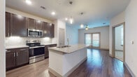 This well-appointed single-floor residence showcases a thoughtfully designed open-concept layout that seamlessly connects the kitchen, dining, and living areas. The kitchen is a standout feature, equipped with stainless steel appliances including a range, microwave, and refrigerator, complemented by rich dark-espresso cabinetry and a classic white subway tile backsplash. A large central island with granite countertops and an integrated sink serves as both a functional workspace and a natural gathering point, illuminated by stylish pendant lights overhead. The living area extends naturally from the kitchen, bathed in natural light from double French-style doors and finished with warm hardwood-style flooring. A ceiling fan adds comfort to the open living space. The bedroom features plush carpeting and a bright window that brings in ample natural light, providing a calm and restful atmosphere. The bathroom is finished with the same high-quality dark cabinetry and granite countertops seen throughout the home, paired with hardwood-style tile flooring, a bathtub, and a walk-in closet. Recessed lighting and consistent modern finishes tie the entire space together, creating a cohesive and contemporary aesthetic throughout.