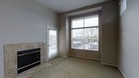 This single-floor space offers a comfortable and functional layout designed with both style and practicality in mind. The living area serves as the focal point of the space, characterized by neutral taupe walls and soft wall-to-wall carpeting that lend a warm, inviting ambiance throughout.

A standout feature of the living area is the tile-surround fireplace, which anchors one wall and adds a classic, cozy element to the room. Adjacent to the fireplace, a glazed exterior door provides convenient direct access to the outdoors and allows additional natural light to filter in.

Opposite the fireplace, a large picture window dominates the wall, framed with clean white trim and fitted with horizontal blinds for privacy control. The oversized window draws in generous natural light and offers an open view of the surrounding outdoor environment, including mature trees and the exterior surroundings.

The elevated placement of the picture window, combined with the generous ceiling height, creates a sense of openness and volume within the living space. The overall design palette — muted tones, clean lines, and understated finishes — provides a versatile backdrop well-suited to a variety of interior styles. This space balances comfort and practicality, making it an approachable and welcoming environment.