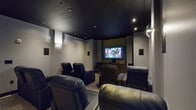 This private home theater is thoughtfully designed to deliver an immersive cinematic experience. The room features a deep navy/dark ceiling that absorbs light and enhances contrast, while the surrounding walls are finished in a cool grey tone, creating a focused, distraction-free viewing environment. Recessed ceiling spotlights provide subtle, ambient illumination that complements the theater atmosphere without interfering with screen visibility.

At the heart of the room is a large wall-mounted flat-screen display, positioned centrally for optimal sightlines from every seat. Beneath the screen sits a handsome dark wood media console with distinctive gold-toned hardware, providing an elegant anchor to the viewing wall. A multi-channel surround sound system is integrated throughout, with wall-mounted speakers positioned on both sides of the room to deliver full, enveloping audio.

Seating consists of multiple rows of plush, dark leather power recliner chairs, each equipped with built-in cup holders — a true nod to the cinema experience. The chairs are arranged to ensure clear views of the screen from every position. The floor is finished with a neutral, textured carpet that adds warmth and aids in sound absorption. A solid dark-stained door provides private entry to the space, completing this well-appointed, dedicated entertainment room.