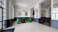 This single-floor space masterfully combines historic architectural grandeur with contemporary interior design. The entrance is defined by soaring arched Gothic-style windows framed in brick, flooding the polished terrazzo floors with abundant natural light and offering clear views to the outdoors. Ornate plaster crown molding, decorative column capitals, and exposed ceiling beams reinforce the building's classic character throughout.

The interior is divided into distinct seating zones, each with its own personality. A central lounge area features a bold emerald-green curved velvet sofa paired with veined marble-topped tables and small crimson ottomans, anchored beneath warm gold chandeliers. Adjacent, a separate room showcases a striking oversized turquoise velvet ottoman as its centerpiece, beneath another dramatic chandelier, complemented by dark wainscoting and gold-framed figurative artwork. Further into the space, a deep magenta curved sectional sofa anchors a secondary lounge, with navy blue seating visible in a more formal dining alcove beyond.

Dark navy wainscoting panels line the lower walls throughout, creating a strong contrast against the crisp white upper walls. The transition from terrazzo near the entrance to rich hardwood flooring deeper in the space adds textural variety, contributing to an overall ambiance that is refined, eclectic, and visually dynamic.