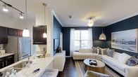 This single-floor residence in Frisco showcases a thoughtfully designed open-concept layout that connects the kitchen and living areas in a cohesive, contemporary arrangement. The kitchen features light granite countertops that contrast elegantly with dark cabinetry, complemented by pendant lighting that adds warmth and definition to the space. A breakfast bar provides a natural transition into the living area, enhancing the open feel.

The living room is anchored by a striking navy blue accent wall that creates a bold focal point against crisp white trim and crown molding. Dark hardwood-style flooring runs throughout, grounding the space with a rich, polished aesthetic. A plush curved sectional sofa in neutral tones pairs with a round wood-and-metal coffee table, offering a balanced mix of comfort and style. Natural light filters in through large windows dressed with white curtains, brightening the room and offering views of the outdoors. Overhead, a decorative ceiling fan and a woven pendant light provide layered illumination. The overall ambiance is modern and inviting, with a curated blend of contrast, texture, and light.