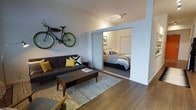 This thoughtfully designed single-floor residence offers a compact yet functional layout that maximizes every square foot. The open-plan living area serves as the heart of the space, featuring rich hardwood-style flooring that runs throughout and creates a warm, cohesive aesthetic. Clean white walls and track lighting overhead contribute to a bright, modern ambiance.

The living area is well-appointed, providing ample room for a full sofa and coffee table arrangement, with a unique wall-mounted bicycle rack serving as a functional design statement. A stylish accent wall treatment and neutral tones accented with pops of yellow add personality and visual interest to the room.

A standout architectural feature is the sliding glass partition that elegantly separates the living area from the bedroom, allowing natural light to flow between the two spaces while maintaining privacy. The bedroom is carpeted for comfort and accommodates a generously sized bed along with bedside furnishings and a closet with a sliding door.

The overall design reflects a contemporary urban aesthetic, blending practicality with style. The open connection between rooms, combined with the thoughtful use of materials and lighting, creates a cohesive and inviting living environment well-suited for modern everyday living.