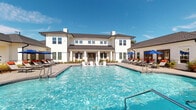 Located at 3215 Midvale Dr in Wilmington, North Carolina, this beautifully appointed community amenity space delivers a resort-like experience designed for relaxation and socializing. The centerpiece is a generously sized swimming pool with crystal-clear blue water, surrounded by a paver pool deck lined with lounge chairs and shade umbrellas, perfect for enjoying the Carolina sunshine. The elegant two-story clubhouse building, clad in crisp white siding with a dark pitched roof, serves as a striking architectural backdrop to the pool area. A spacious covered pavilion anchors one end of the outdoor space, featuring a wood-paneled ceiling supported by white columns. This pavilion is well-equipped with an outdoor fireplace framed in white brick with a herringbone tile surround, comfortable wicker lounge seating with bold accent cushions, dining tables and chairs, and a built-in grilling and bar station ideal for gatherings. The pavilion opens to sweeping views of the surrounding green spaces and residential community beyond the deck railing. A separate spa and pool house structure provide additional convenience. The overall ambiance is welcoming and upscale, blending classic coastal design with modern outdoor living amenities.
