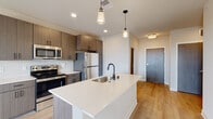 This single-floor unit showcases a thoughtfully designed open-concept layout with contemporary finishes. The kitchen serves as a central focal point, featuring a large island with a white quartz countertop, an undermount sink, and a matte black faucet. Pendant lights hang above the island, complementing the recessed ceiling lighting throughout. Dark wood-grain cabinetry extends to the ceiling, providing generous storage, while stainless steel appliances—including a refrigerator, range, and microwave—complete the functional cooking space. Light wood-tone flooring runs through the kitchen and living area, lending warmth to the open layout. Sliding glass doors lead to a private balcony, allowing natural light to fill the living space.

The bedrooms feature plush carpeting and neutral walls, creating a calm and comfortable atmosphere. One bedroom benefits from a large window with outdoor views and a ceiling fan, along with a closet finished in warm wood tones. The bathroom is appointed with a dark-framed vanity mirror, quartz countertop, undermount sink, modern fixtures, and a shower-tub combination with sliding glass doors. In-unit laundry is also present, adding everyday convenience to this well-appointed space.