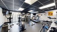 Located at 18645 Detroit Ave in Lakewood, Ohio, this fitness center offers a comprehensive, single-floor workout facility designed to support a wide range of exercise routines. The space features a dark drop-tile ceiling with recessed panel lighting that keeps the room bright and energized, while large mirrors line the walls to enhance the sense of space and allow for proper form checks during exercise.

The cardio section is well-appointed with multiple treadmills, elliptical trainers, and a stationary spin bike, providing ample options for aerobic training. A dedicated free weights area includes a full dumbbell rack with a wide range of weight increments, complemented by an adjustable workout bench for strength training. A multi-function cable machine anchors the strength-training zone, enabling a broad variety of resistance exercises.

Additional equipment includes a rowing machine, medicine balls mounted on a wall rack, a stability ball, and a yoga/stretching mat area for flexibility and core work. Motivational fitness artwork decorates the white walls, contributing to an encouraging atmosphere. Rubber flooring throughout the space provides durability, safety, and comfort underfoot. A wall-mounted television is also present, allowing users to stay entertained during workouts. Natural light filters in through vertical-blind windows, rounding out this functional and well-maintained fitness facility.
