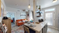 This single-floor residence offers a thoughtfully arranged layout that balances function and style throughout. The open-concept living and dining area serves as the heart of the home, where light wood-look flooring, neutral walls, and large windows with blinds create a bright, welcoming atmosphere. A pendant light fixture anchors the dining space adjacent to the kitchen counter, which doubles as a breakfast bar. A seating nook at the far end of the living area provides an additional area for relaxation, bathed in natural light from multiple windows.

The galley-style kitchen is efficiently designed with white upper cabinetry and contrasting gray lower cabinets, all topped with quartz-style countertops. Stainless steel appliances include a refrigerator, dishwasher, microwave, and range. Open floating shelves mounted on one wall offer display and storage space, complemented by a direct exterior door that brings in additional natural light.

The bedroom is a calm retreat featuring dual windows, a stylish sliding barn door that opens to a walk-in closet with built-in storage. The bathroom includes a full tub-shower combination with clean white tile surround and chrome fixtures, maintaining the unit's cohesive, modern aesthetic throughout.