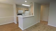 This single-floor residence offers a practical and well-organized layout with a clean, neutral aesthetic throughout. The kitchen is the focal point of the main living area, designed in an open-concept style that connects seamlessly to the adjacent living space. Light wood cabinetry provides ample storage, while white appliances — including a refrigerator, electric range, and over-the-range microwave — offer full functionality. A peninsula counter with a white surface extends into the living area, creating a natural divide between the kitchen and the open floor plan beyond, while also serving as a casual dining or prep space. Recessed and overhead lighting keeps the kitchen bright and well-illuminated. The living area features light hardwood-style flooring that transitions to carpet, giving each zone a distinct feel while maintaining a cohesive overall look. Soft gray walls run throughout, contributing to a calm and neutral atmosphere. White trim and baseboards add a crisp finishing detail to every room. A hallway visible from the main living area suggests additional rooms beyond, completing this thoughtfully designed single-level home.