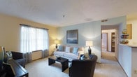 Located at 4800 Lake Forest Blvd in Westerville, Ohio, this single-floor residence offers a thoughtfully designed layout with a warm and inviting ambiance throughout. The living room serves as the centerpiece of the home, featuring plush wall-to-wall carpeting that adds comfort underfoot. Soft, neutral wall tones are complemented by a calming blue accent wall, creating a serene and stylish atmosphere. Natural light filters in through large windows dressed with sheer curtains and blinds, brightening the space and enhancing its open feel. The living area comfortably accommodates a full seating arrangement, including a sofa and accent chairs, anchored by a sleek coffee table. Overhead, a textured ceiling adds subtle architectural character, while recessed ventilation ensures climate comfort. Tasteful wall art and coordinated décor contribute to a cohesive aesthetic. An open sightline connects the living room to adjacent areas of the home, suggesting a functional and accessible floor plan. The overall design balances practicality with comfort, making this Westerville residence a well-rounded living space suited for everyday living.