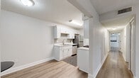This single-floor residence offers a clean, modern aesthetic defined by light gray walls, luxury vinyl plank flooring, and textured ceilings throughout. The layout includes multiple living and sleeping areas connected by a well-organized corridor, as seen in the 3D model overview.

Two distinct kitchen configurations are present: one is a galley-style kitchen lined with white shaker cabinets, quartz-style countertops, a subway tile backsplash, and a full suite of stainless steel appliances including a gas range, dishwasher, refrigerator, and over-the-range microwave. A second, more open kitchen area features similar white cabinetry, a stainless steel sink, and additional appliances, partially separated from adjacent space by a structural column.

A dedicated dining nook provides an intimate space for meals, while the living area offers a ceiling fan and access to a private balcony with outdoor seating and tree views through sliding glass doors. Bedrooms are generously sized with ceiling fans, closets, and natural light. One room is configured as a home office with a window overlooking greenery.

The bathroom features a white vanity with a marble-style countertop, a large wall mirror, and a tub-shower combination. In-unit laundry with a washer and dryer completes the practical amenities of this well-appointed space.