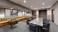 Located at 608 Second Ave S in downtown Minneapolis, this thoughtfully designed amenity space offers a versatile environment suited for both collaborative work and relaxed social gatherings. The layout encompasses a primary lounge and meeting room connected by two well-lit hallways, creating a cohesive and navigable flow throughout.

The main room serves as the centerpiece, featuring a large conference-style table surrounded by sleek, dark barrel chairs with brass-accented legs — ideal for meetings or group work sessions. Along the perimeter, a cushioned tan banquette runs the length of one wall, complemented by tufted green lounge chairs and dark marble-topped tables, providing a more casual seating alternative. A built-in wood bookcase stocked with books and décor adds warmth and a residential feel, while a recessed nook with upholstered bench seating offers a semi-private retreat.

The walls are finished with a subtle grid-patterned tile, lending texture and a refined, contemporary aesthetic throughout. Recessed ceiling lighting and wall sconces provide warm, layered illumination. Framed black-and-white photography adorns the hallways, adding character to the transition spaces. The corridors feature wood-framed doors with sidelights, gray carpet, and clean wall finishes, ensuring a polished and professional atmosphere from entry to exit.
