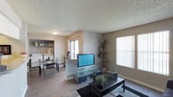 This single-floor residence offers a comfortable and functional layout with an open-concept design that seamlessly connects the living and dining areas. The living room is bright and welcoming, benefiting from multiple windows dressed with vertical blinds that allow generous natural light to fill the space. Durable laminate flooring runs throughout, providing a clean and cohesive look, while a textured popcorn ceiling adds a classic finishing touch. A partial wall divider subtly defines the transition between the living and dining zones without compromising the open feel of the interior. The dining area features a built-in shelving unit, offering both display and storage functionality, and receives its own natural light through an adjacent window. The overall interior palette is neutral and understated, creating a versatile backdrop. Beyond the unit, the outdoor common area is a standout feature, showcasing a landscape of tall, mature pine and palm trees that provide ample shade over a spacious grounds area. Manicured shrubs, open pathways, and what appears to be a recreational court in the background round out the exterior amenities, offering residents a pleasant and green outdoor environment to enjoy.