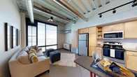 This single-floor space showcases a striking industrial-modern aesthetic that blends raw architectural elements with contemporary comfort. The open-plan layout seamlessly connects the living area and kitchen, creating a cohesive and functional environment. Exposed silver ductwork and ventilation systems run across the ceiling, complemented by track lighting and a ceiling fan, lending the space an urban loft character. Soaring ceilings amplify the sense of openness throughout.

The living area is anchored by large floor-to-ceiling windows that flood the space with abundant natural light and offer expansive exterior views. The kitchen is well-appointed with light maple cabinetry, a stainless steel refrigerator, a gas range, a microwave, and ample shelving for storage. A breakfast bar provides a casual dining option and serves as a natural divider between the kitchen and living zones.

The flooring features a combination of carpet in the living area and hard flooring near the kitchen, adding visual contrast and practical zoning. The overall ambiance is bright, airy, and contemporary, with the interplay of industrial details and warm finishes creating an inviting yet stylish atmosphere well-suited to modern urban living.