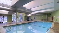 This indoor pool facility is designed to offer a bright, welcoming aquatic environment. The space is anchored by a generously sized rectangular pool with clear, blue-tinted water that reflects the surrounding architecture. The ceiling features prominent skylights that flood the area with natural light, complementing the large arched windows along one wall, which offer views of the outdoors and further enhance the airy atmosphere. Structural columns are positioned throughout, adding to the architectural character of the space. The walls are painted in a soft sage green, contributing to a calm and refreshing ambiance. A wide tiled pool deck surrounds the water on multiple sides, furnished with lounge chairs and seating areas, providing ample space for relaxation. Access to the outdoors is available through glass-panel doors, connecting the interior to an exterior area. The overall design balances functionality with comfort, making it a well-appointed facility suitable for both leisure and fitness use.