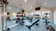 This fitness center is designed to support a full range of workout routines in a bright, open environment. The space features vaulted ceilings that lend an airy, expansive feel, while large windows along multiple walls flood the interior with natural light and offer views of the surrounding greenery outdoors. Light-toned vinyl plank flooring runs throughout, contributing to the clean, modern aesthetic.

The facility is thoroughly equipped for both cardio and strength training. Cardio machines include treadmills, an elliptical trainer, and multiple spin bikes, all positioned to take advantage of the natural light from the large windows. For strength training, the space houses a cable machine system suitable for a wide variety of resistance exercises, an adjustable weight bench, a dumbbell rack with multiple weight options, and a well-stocked medicine ball rack with a colorful array of weighted balls for functional fitness training.

Full-length mirrors are installed along one wall, enhancing the sense of space and allowing users to monitor their form during workouts. Ceiling fans with pendant-style fixtures provide ventilation and comfort, while recessed lighting supplements natural light throughout the day. Multiple flat-screen televisions are mounted on the walls, offering entertainment during workouts. The overall ambiance is clean, well-maintained, and energizing, making it a functional and inviting space for fitness.