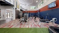 Located at 1335 Herrington Rd in Duluth, GA, this fitness center is a purpose-built facility designed to support a wide range of training styles. The space is defined by its striking interior design, featuring bold navy and red two-tone walls that create an energetic, athletic atmosphere throughout.

The facility is divided into two distinct training zones. The strength and equipment area features patterned carpet tiles in complementary red and neutral tones, and is outfitted with a full smith machine, a comprehensive dumbbell rack, multiple cable and resistance machines, treadmills, air bikes, and wall-mounted televisions for entertainment during workouts. Colorful artwork accents the walls, adding visual interest to the space.

Adjacent to this is a generous functional training zone covered in synthetic turf, ideal for dynamic and high-intensity workouts. This area includes a multi-functional rig with suspension trainers, battle ropes, a heavy bag, weighted sleds, and oversized tires for functional fitness drills.

Soaring ceilings with exposed beam detailing and a mezzanine-level railing above add architectural depth to the space. An abundance of large clerestory windows allows natural light to flood both zones, with views of surrounding trees enhancing the bright and motivating environment.
