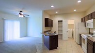 This single-floor residence offers an open-concept design that seamlessly connects the kitchen and living areas, creating a cohesive and functional layout. The kitchen is anchored by a peninsula with a double-basin sink and granite-style countertops, complemented by rich dark cabinetry above and recessed lighting throughout. The transition between the tiled kitchen floor and the carpeted living area is smooth, with the living space featuring a ceiling fan overhead. Adjacent to the kitchen, a built-in shelving unit provides convenient open storage, ideal for organization and display. A dedicated laundry area is tucked nearby, housing side-by-side washer and dryer units behind a paneled door, adding significant practicality to the space. The overall color palette is neutral, with warm beige walls and light-toned tile flooring in the main areas, contributing to a bright and welcoming ambiance. Recessed lighting ensures even illumination throughout, enhancing the open feel of the interior. This well-appointed space balances everyday functionality with a clean, understated aesthetic.