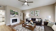 This thoughtfully designed single-floor residence offers a cohesive layout connecting comfortable living spaces with a warm, neutral aesthetic accented by green tones throughout. The living room serves as a central gathering area, highlighted by a white-framed fireplace, rich hardwood-style flooring, a ceiling fan, and ample wall space showcasing bold modern artwork. Large windows in the living area invite natural light and provide a view to an outdoor patio, enhancing the sense of openness. Adjacent to the living room, a dedicated dining area features hardwood-style flooring, wall mirrors that amplify the space, and room for a full-size dining table. The kitchen is equipped with white cabinetry, stainless steel appliances including a refrigerator and dishwasher, a double sink, and clean tile flooring. The primary bedroom offers plush carpet, a ceiling fan, a closet, and generous space for a large bed and furniture suite. A second bedroom provides similar comfort with soft carpet and access to natural light. The primary bathroom features a wide dual-sink vanity with a large mirror, ample under-counter storage, and tile flooring, while the second bathroom offers a single-sink vanity with a tub and shower combination.