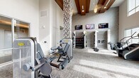 Located at 6611 E Mayo Blvd in Phoenix, AZ, this fitness center is a well-appointed, single-floor facility designed to support a comprehensive workout experience. The space is defined by its dramatic soaring ceilings accented with rich wood beam detailing, floor-to-ceiling windows that flood the interior with natural light, and views of the surrounding landscaped grounds.

The main workout floor is generously proportioned and houses an extensive row of treadmills and cardio machines positioned along the large windows, offering an energizing outdoor view during workouts. Strength training is well represented with a broad selection of Technogym selectorized machines, a functional training rig, cable systems, and a fully stocked free weight area complete with dumbbells and medicine balls organized along a dedicated wall-mounted rack. Multiple flat-screen televisions are mounted throughout, keeping members engaged during their sessions.

A standout architectural feature is the mosaic-tiled columns that anchor the space, adding a sophisticated design element to the otherwise clean, neutral palette of gray tones and textured wall finishes. A separate enclosed studio room with floor-to-ceiling glass walls and mirrored surfaces provides a dedicated space for group fitness, stretching, or personal training. Additional amenities include open shelving, storage areas, and a Concept2 rowing machine. The patterned carpet throughout ties the space together with a modern, polished aesthetic.