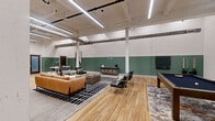 Located at 201 S 12th St in Bessemer City, NC, this single-floor community space offers a generously sized, open-plan layout that masterfully blends industrial character with modern design. Soaring ceilings with exposed ductwork and structural columns pay homage to the building's industrial roots, while sleek linear LED lighting illuminates the entire space with a clean, contemporary glow. The walls feature a distinctive two-tone treatment — crisp white upper halves contrasted by a rich sage green lower half — lending a cohesive and inviting aesthetic throughout.

Warm-toned hardwood flooring flows seamlessly across the space, anchoring multiple distinct zones. A central lounge area is outfitted with a tan leather sofa, accent chairs, and a bold geometric area rug, creating a comfortable gathering focal point. Adjacent to it, a billiards area centers around a full-size pool table, complemented by wall-mounted cue storage and casual seating. A kitchenette at the rear features stainless steel appliances, ample cabinetry, a bar-style counter, and pendant lighting, making it well-suited for events and social gatherings.

A dining area with a marble-top table and modern chairs, curated artwork on the walls, and a large glass pass-through window connecting to an adjacent corridor round out this thoughtfully designed, multifunctional space.