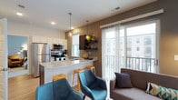 Located at 8900 Lyra Dr in Columbus, Ohio, this single-floor residence offers a thoughtfully designed open-concept layout that seamlessly connects the kitchen and living areas. The kitchen is appointed with stainless steel appliances, including a refrigerator, microwave, and range, complemented by crisp white cabinetry and a light stone countertop island that doubles as a breakfast bar with bar seating. Pendant lighting hangs above the island, adding a modern, polished touch to the space. Dark floating shelves mounted on a contrasting accent wall provide stylish display and storage options. The living area flows naturally from the kitchen, creating a cohesive and functional gathering space. Large floor-to-ceiling sliding glass doors flood the interior with abundant natural light and open to a private balcony, offering views of the surrounding urban landscape. The neutral color palette, featuring warm taupe and white tones, is accented by contemporary furnishings, contributing to a clean and inviting ambiance. The light hardwood-style flooring ties the open space together, enhancing the bright and airy atmosphere throughout this well-appointed Columbus residence.