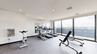 The fitness center at 2501 Oakford St in Philadelphia's 19146 neighborhood is a thoughtfully designed workout space that combines modern amenities with an open, light-filled environment. The room features a clean, contemporary aesthetic characterized by crisp white walls, recessed lighting, and durable rubber flooring suited for high-intensity exercise.

Large floor-to-ceiling windows along one wall flood the space with natural light while offering sweeping views of the Philadelphia skyline and surrounding neighborhood, creating an energizing atmosphere for workouts. A full-length glass partition and strategically placed wall mirrors further enhance the sense of openness and allow users to monitor their form during exercise.

The cardio zone is well-appointed, featuring multiple treadmills and an elliptical trainer lined along the mirrored wall. The strength training area includes a multi-functional cable machine, adjustable weight benches, a dedicated cycling bike, and a rowing machine. A tiered dumbbell rack stocked with a range of free weights, along with a barbell rack and medicine balls, rounds out the available equipment.

The space also benefits from an adjacent outdoor area visible through the large sliding glass doors, contributing to the bright and airy ambiance. Overall, the fitness center offers a comprehensive and inviting environment for a full range of workout routines.