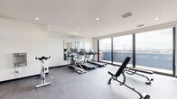 The fitness center at 2501 Oakford St in Philadelphia's 19146 neighborhood is a thoughtfully designed workout space that combines modern amenities with an open, light-filled environment. The room features a clean, contemporary aesthetic characterized by crisp white walls, recessed lighting, and durable rubber flooring suited for high-intensity exercise.

Large floor-to-ceiling windows along one wall flood the space with natural light while offering sweeping views of the Philadelphia skyline and surrounding neighborhood, creating an energizing atmosphere for workouts. A full-length glass partition and strategically placed wall mirrors further enhance the sense of openness and allow users to monitor their form during exercise.

The cardio zone is well-appointed, featuring multiple treadmills and an elliptical trainer lined along the mirrored wall. The strength training area includes a multi-functional cable machine, adjustable weight benches, a dedicated cycling bike, and a rowing machine. A tiered dumbbell rack stocked with a range of free weights, along with a barbell rack and medicine balls, rounds out the available equipment.

The space also benefits from an adjacent outdoor area visible through the large sliding glass doors, contributing to the bright and airy ambiance. Overall, the fitness center offers a comprehensive and inviting environment for a full range of workout routines.