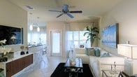 This charming single-floor residence showcases a coastal-inspired aesthetic with a light, airy atmosphere throughout. The open-concept layout seamlessly connects the living and dining areas, creating a sense of spaciousness and ease of flow. The living room is elegantly appointed with a plush white sectional sofa accented by teal and patterned throw pillows, anchored by a sleek glass coffee table resting on a bold black area rug. A ceiling fan overhead adds both function and a relaxed, breezy feel to the space. Large windows dressed with sheer white curtains invite abundant natural light, while a vibrant tropical plant and coastal-themed wall art reinforce the serene beach-inspired ambiance. The adjacent kitchen and dining area feature a modern breakfast bar with contemporary white bar stools and stylish pendant lighting, complemented by dark countertops that provide a striking contrast to the otherwise bright palette. Light-toned tile flooring extends throughout, enhancing the open and clean character of the home. Overall, this space offers a welcoming and well-curated environment that balances comfort with sophisticated coastal style.