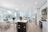 This contemporary high-rise residence is defined by clean lines, a bright neutral palette, and abundant natural light streaming through expansive floor-to-ceiling windows. The open-plan living, dining, and kitchen area forms the heart of the home, seamlessly connecting the main living spaces while offering panoramic city skyline views from multiple angles. A covered balcony extends the living area outdoors, providing an ideal vantage point over the urban landscape.

The kitchen is thoughtfully designed with flat-front cabinetry in white and warm grey tones, a large central island with a quartz countertop and built-in wine rack, stainless steel appliances including a microwave, oven, and full-size refrigerator, and bar seating for casual dining. Light wood flooring flows throughout the main living areas, enhancing the sense of warmth and continuity.

The living room is bathed in natural light from oversized windows on two sides, framing striking skyline vistas. Two well-proportioned bedrooms each feature large windows with city views and light wood flooring. Two bathrooms serve the residence — one with a soaking tub and tiled surround, and the other with a walk-in shower — both finished in clean white tones with modern fixtures. An entry area with a frosted glass door completes the well-appointed layout.