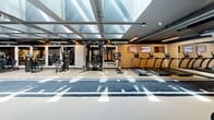 Located in the heart of Miami's vibrant Wynwood arts district, WYND27 is a purpose-built fitness facility offering a comprehensive and well-equipped exercise environment. The single-floor space is defined by its striking architectural ceiling — a large skylight grid that floods the main floor with natural light, creating an open and energizing atmosphere throughout the day. A branded turf sprint track runs the full length of the facility, serving as the central axis around which all equipment is organized, and doubles as a functional training lane for speed and agility work. Along the perimeter and flanking the track, the facility houses an extensive array of strength and conditioning equipment, including cable machines, multi-functional rigs, plate-loaded machines, selectorized weight stations, treadmills, stationary bikes, adjustable benches, and a full dumbbell rack. A dedicated functional training zone features a punching bag, medicine balls, kettlebells, resistance bands, and a large tire for dynamic training. Full-length mirrored walls in select areas enhance the sense of space and allow for form monitoring during workouts. The flooring transitions between rubberized gym surfaces and turf, providing appropriate traction for varied exercises. Linear LED lighting complements the natural daylight, ensuring consistent illumination throughout. The overall design is modern and industrial, with an aesthetic that reflects the creative energy of the surrounding Wynwood neighborhood.