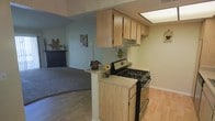 This single-floor residence offers a practical and comfortable layout designed to maximize functionality and flow. The kitchen is outfitted with light oak cabinetry that provides generous storage space, complemented by a neutral countertop surface. A gas range with overhead ventilation serves as the focal point of the cooking area, while a skylight-style ceiling fixture brightens the space with added illumination. The kitchen opens seamlessly to the living area via a half-wall peninsula, which doubles as a convenient prep and serving surface, adorned with decorative touches that add a homey character. The transition between the kitchen and living room is marked by a shift from light hardwood flooring to plush carpeting, creating a natural delineation between the two spaces. The living room features a classic fireplace with a dark surround, serving as a warm and inviting anchor to the room. Soft blue-green wall tones carry throughout, lending the interior a calm, cohesive ambiance. The single-level layout ensures easy accessibility and a connected feel throughout the home, making it well-suited for everyday living.