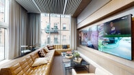 This single-floor lounge is defined by its contemporary architectural design and carefully curated interiors. Expansive floor-to-ceiling windows wrap around the space, flooding it with natural light and framing views of the surrounding urban brick buildings. A linear slatted ceiling treatment adds visual depth and a refined, modern character throughout.

The main seating area centers on a generously sized tufted caramel leather sectional sofa, complemented by sculptural white accent chairs with curved forms and patterned upholstery. A sleek black marble-topped coffee table anchors the arrangement, styled with decorative objects and greenery. On the opposing wall, a built-in wood-paneled feature wall houses a large multi-panel LG display configuration alongside integrated shelving with dark stone accents, creating a polished media-focused focal point.

Floor-length drapery in soft grey tones lines the window walls, offering flexible light control. A compact bistro-style seating nook with a marble-topped brass-legged table and a sculptural white chair provides an intimate corner for smaller gatherings. Glass doors open directly to an outdoor terrace furnished with seating, extending the usable area outdoors. An adjacent entrance leads to additional amenity spaces, indicating this lounge is part of a larger facility.